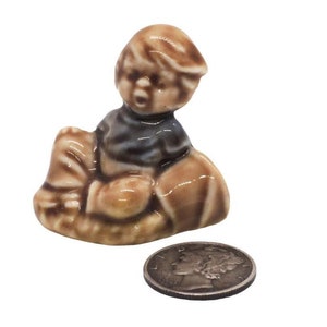May include: A ceramic figurine of a boy sitting on a brown and white base. The boy is wearing a blue and white shirt and has brown hair.
