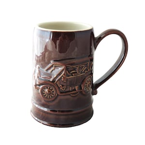 May include: A brown ceramic mug with a raised design of a vintage car. The mug has a white interior and a handle.