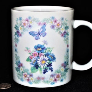 K&ouml;nnte beinhalten: Wei&szlig;e Keramiktasse mit einem Blumendesign mit blauen, rosa und violetten Blumen, gr&uuml;nen Bl&auml;ttern und einem blauen Schmetterling. Die Tasse hat einen wei&szlig;en Henkel.