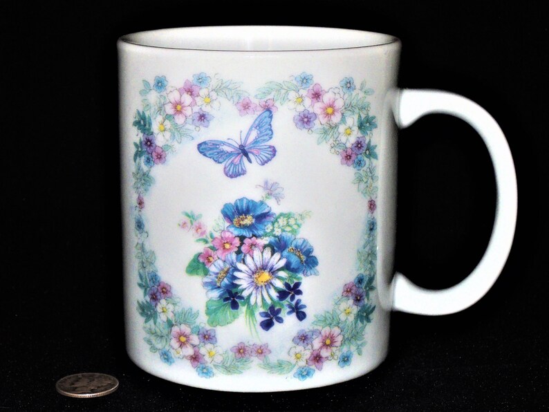 K&ouml;nnte beinhalten: Wei&szlig;e Keramiktasse mit einem Blumendesign mit blauen, rosa und violetten Blumen, gr&uuml;nen Bl&auml;ttern und einem blauen Schmetterling. Die Tasse hat einen wei&szlig;en Henkel.