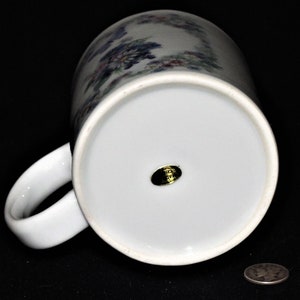 K&ouml;nnte beinhalten: Wei&szlig;e Keramiktasse mit einem Blumendesign in Blaut&ouml;nen, Rosa und Gr&uuml;n. Die Tasse hat einen goldenen Aufkleber auf der Unterseite mit dem Text "Made in Japan".