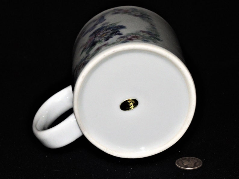 K&ouml;nnte beinhalten: Wei&szlig;e Keramiktasse mit einem Blumendesign in Blaut&ouml;nen, Rosa und Gr&uuml;n. Die Tasse hat einen goldenen Aufkleber auf der Unterseite mit dem Text "Made in Japan".