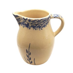 May include: A tan ceramic pitcher with a curved handle and a spout. The rim and handle are decorated with a blue floral pattern. A blue floral design is also painted on the front of the pitcher.