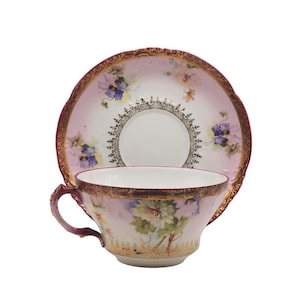 K&ouml;nnte beinhalten: Ein Vintage-Tee-Set, bestehend aus einer Tasse und einer Untertasse. Die Tasse und die Untertasse sind mit einem rosa-wei&szlig;en Blumendesign mit goldenen Verzierungen bemalt. Die Untertasse hat einen gewellten Rand.