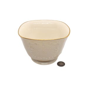 May include: A square, ivory-colored porcelain bowl with a gold rim and a delicate embossed pattern. The bowl is sitting on a white surface.