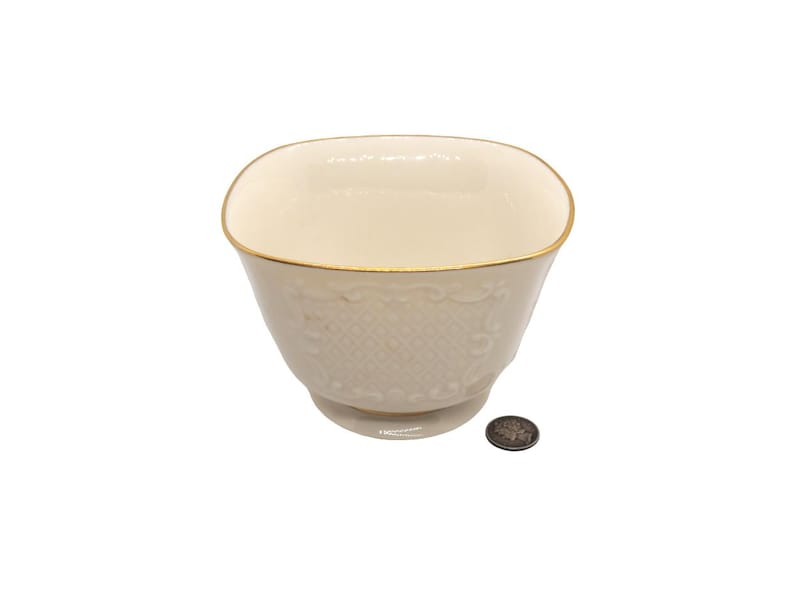 May include: A square, ivory-colored porcelain bowl with a gold rim and a delicate embossed pattern. The bowl is sitting on a white surface.