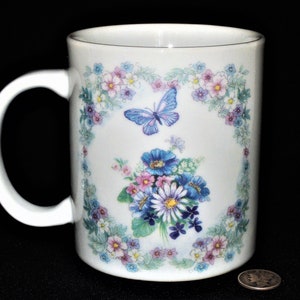 K&ouml;nnte beinhalten: Wei&szlig;e Keramiktasse mit einem Blumendesign mit blauen und rosa Bl&uuml;ten, gr&uuml;nen Bl&auml;ttern und einem blauen Schmetterling. Das Design ist von einem Rand aus kleineren Blumen eingerahmt.