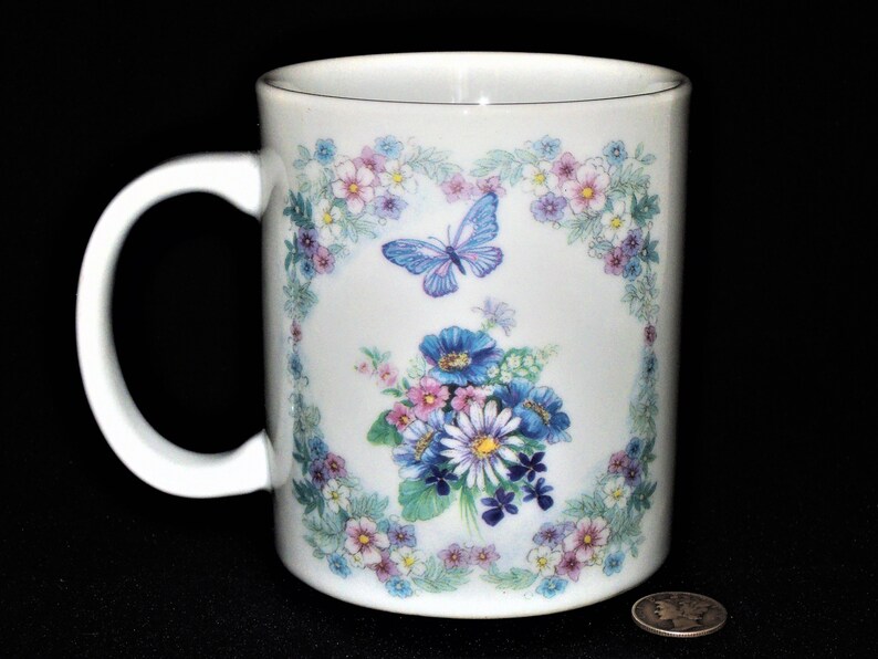 K&ouml;nnte beinhalten: Wei&szlig;e Keramiktasse mit einem Blumendesign mit blauen und rosa Bl&uuml;ten, gr&uuml;nen Bl&auml;ttern und einem blauen Schmetterling. Das Design ist von einem Rand aus kleineren Blumen eingerahmt.