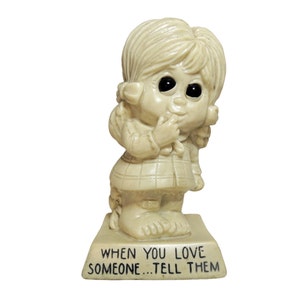 May include: A white ceramic figurine of a girl with large eyes and pigtails. The figurine is standing on a base with the inscription "When you love someone...tell them".