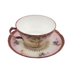 K&ouml;nnte beinhalten: Ein Vintage-Tee-Set, bestehend aus einer Tasse und einer Untertasse. Die Tasse und die Untertasse sind haupts&auml;chlich rosa mit floralen und goldenen Akzenten. Der Rand der Tasse und der Untertasse ist tiefrot.
