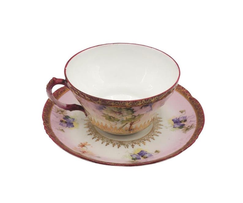K&ouml;nnte beinhalten: Ein Vintage-Tee-Set, bestehend aus einer Tasse und einer Untertasse. Die Tasse und die Untertasse sind haupts&auml;chlich rosa mit floralen und goldenen Akzenten. Der Rand der Tasse und der Untertasse ist tiefrot.