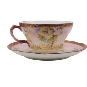 K&ouml;nnte beinhalten: Ein Vintage-Tee-Set, bestehend aus einer Tasse und einer Untertasse. Die Tasse und die Untertasse sind hellrosa mit einem Blumendesign. Der Rand der Tasse und der Untertasse ist mit Gold verziert. Der Henkel der Tasse ist dunkelrosa.