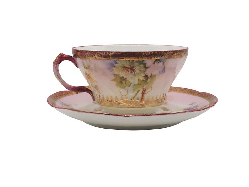 K&ouml;nnte beinhalten: Ein Vintage-Tee-Set, bestehend aus einer Tasse und einer Untertasse. Die Tasse und die Untertasse sind hellrosa mit einem Blumendesign. Der Rand der Tasse und der Untertasse ist mit Gold verziert. Der Henkel der Tasse ist dunkelrosa.