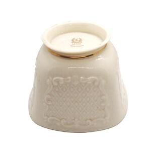 May include: A white ceramic cup with a gold rim and intricate embossed design. The cup has a raised, embossed pattern of diamonds and swirls. The bottom of the cup has a gold rim and the words "Lenox" and "Made in USA" are printed in gold.