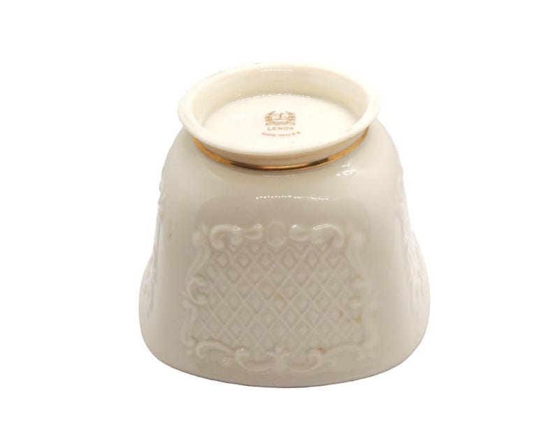 May include: A white ceramic cup with a gold rim and intricate embossed design. The cup has a raised, embossed pattern of diamonds and swirls. The bottom of the cup has a gold rim and the words "Lenox" and "Made in USA" are printed in gold.