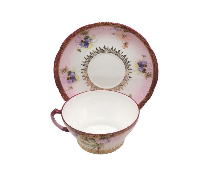 K&ouml;nnte beinhalten: Vintage Tee-Set, bestehend aus Tasse und Untertasse. Die Tasse und Untertasse haben einen rosa-wei&szlig;en Hintergrund mit floralen und goldenen Akzenten. Der Rand der Tasse und Untertasse ist goldfarben.