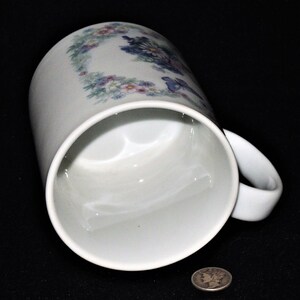 K&ouml;nnte beinhalten: Wei&szlig;e Keramiktasse mit einem Blumendesign in Blaut&ouml;nen, Rosa und Gr&uuml;n. Die Tasse hat einen Henkel und ist leicht zur Seite geneigt.