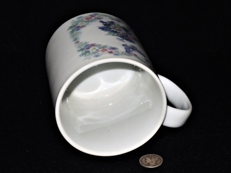 K&ouml;nnte beinhalten: Wei&szlig;e Keramiktasse mit einem Blumendesign in Blaut&ouml;nen, Rosa und Gr&uuml;n. Die Tasse hat einen Henkel und ist leicht zur Seite geneigt.