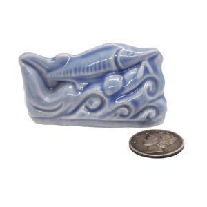 May include: A blue ceramic figurine of a sturgeon fish swimming in waves. The figurine is about the size of a silver dollar.