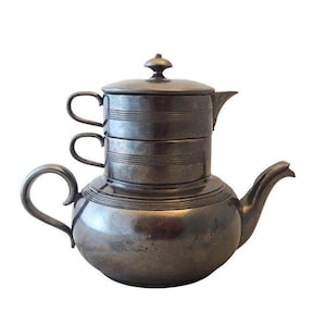 May include: Antique silver-toned tea set with a teapot and two stacked cups. The teapot has a rounded body, a curved spout, and a large handle. The cups have small handles. The set is likely made of metal.