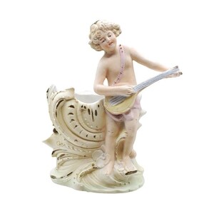 May include: A vintage porcelain figurine of a cherub playing a stringed instrument. The cherub is nude, with a pink strap across the chest. The base is a cream-colored shell-like design with gold accents.