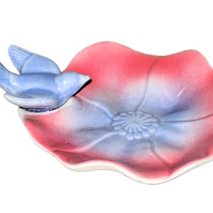 May include: A blue bird perched on a pink and blue ceramic flower dish. The dish is shaped like a flower with a center that has a swirl design.
