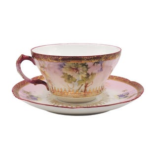 K&ouml;nnte beinhalten: Ein Vintage-Tee-Set. Die Tasse und Untertasse sind rosa mit goldenen Verzierungen und Blumendesigns. Die Tasse hat einen gebogenen Henkel und die Untertasse einen gewellten Rand. Ein klassisches Teeservice.