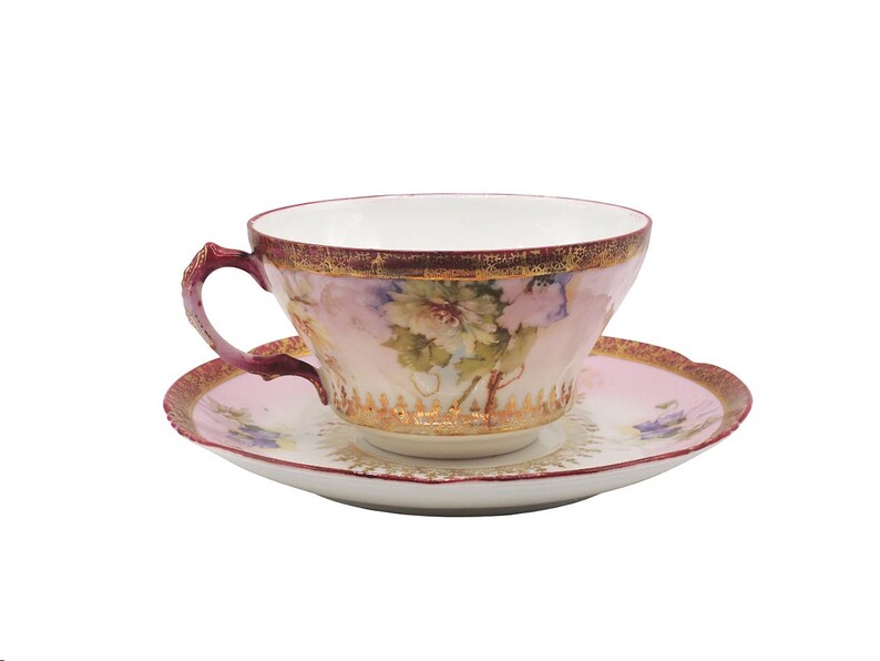 K&ouml;nnte beinhalten: Ein Vintage-Tee-Set. Die Tasse und Untertasse sind rosa mit goldenen Verzierungen und Blumendesigns. Die Tasse hat einen gebogenen Henkel und die Untertasse einen gewellten Rand. Ein klassisches Teeservice.