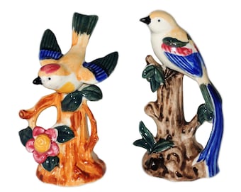 May include: Two ceramic bird figurines perched on tree branches. The bird on the left is yellow, blue, and red with its wings spread. The bird on the right is white, blue, and red with a long tail.