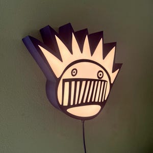 May include: A wall-mounted light fixture shaped like a cartoon face with a spiky crown. The fixture has a black outline with a white interior that illuminates when turned on. The background is a solid green color.