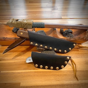 May include: A handcrafted wooden knife with a dark leather sheath, adorned with gold-colored studs. The knife features a wooden handle and a sharp blade. A carved wooden handle with a decorative design is also visible.
