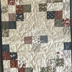 May include: A rectangular table runner featuring a cream-colored fabric base with an intricate, continuous swirling quilted pattern. The edges are decorated with a patchwork design of small squares in shades of navy blue, rust red, and olive green.