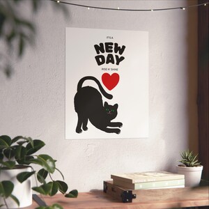 Könnte beinhalten: Ein weißes Poster mit einer schwarzen Katzen-Silhouette, die sich zu einem roten Herzen streckt. Der Text "IT'S A NEW DAY RISE N' SHINE" steht über der Katze. Das Poster befindet sich auf einem Holzregal mit Pflanzen und Büchern.