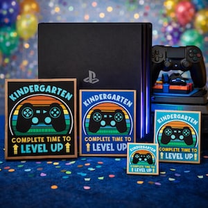 May include: A black PlayStation console with a controller and several wooden signs. The signs read "Kindergarten Complete Time to Level Up" with a controller graphic in a retro color scheme. The signs are in various sizes.