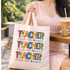 May include: A cream-colored tote bag with the word "TEACHER" in colorful letters, repeated three times. Below each word are the words "teach love inspire". The bag has cream-colored handles.
