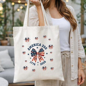 May include: Cream-colored tote bag featuring a patriotic design. The bag displays a large bow with red, white, and blue stripes and stars, with the text "AMERICA 250" and the years "1776" and "2026". Small bow accents surround the central design.