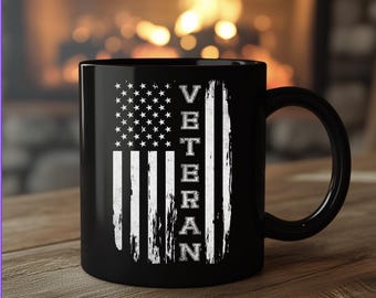 May include: A black ceramic mug featuring a white distressed graphic of the American flag with the word VETERAN printed vertically in a bold, sans-serif font. The design includes a field of stars and vertical stripes on the front of the mug.