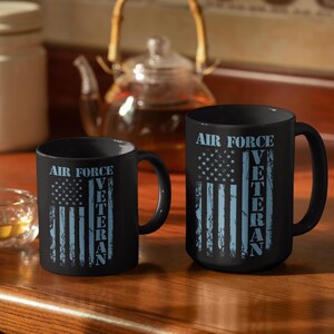 May include: Two black ceramic mugs featuring a light blue graphic of the American flag and the text AIR FORCE VETERAN. The design has a distressed, weathered appearance. The mugs have a standard cylindrical shape with a side handle.