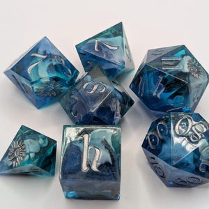 Puede incluir: Un conjunto de siete dados poliédricos en tonos azules, con números y diseños florales plateados. Los dados están hechos de un material translúcido, con un efecto veteado. Dados para juegos de mesa.