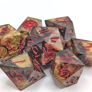 Puede incluir: Un conjunto de siete dados poliédricos con un diseño único. Los dados presentan una mezcla de tonos dorados y marrones con numeración roja. La estética general recuerda a un juego de fantasía o rol.