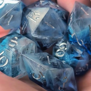 Puede incluir: Un conjunto de siete dados poliédricos con efecto veteado azul y blanco. Los dados tienen numeración plateada y están hechos de resina transparente. Estos dados están diseñados para juegos de rol de mesa.
