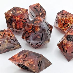 Puede incluir: Juego de siete dados poliédricos con un diseño de hojas doradas y marrón rojizo. Los dados son transparentes con números negros y un diseño floral en un dado. Ideal para juegos de mesa.