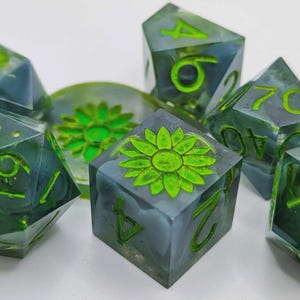 Puede incluir: Un conjunto de dados poliédricos en resina azul-verdosa translúcida, con números verdes y un diseño floral verde. Estos dados están diseñados para juegos de mesa y rol.