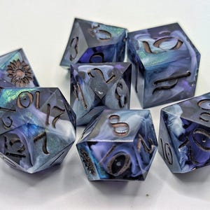 Puede incluir: Un conjunto de siete dados poliédricos con un diseño arremolinado azul, morado y blanco. Los dados tienen números de color bronce y un girasol en un dado. Para juegos de mesa.