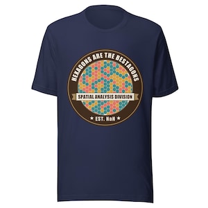 Pode incluir: Camiseta azul marinho com um design circular. O design apresenta um padrão de favo de mel em tons de laranja, amarelo, azul e verde. O texto inclui "HEXAGONS ARE THE BESTAGONS" e "SPATIAL ANALYSIS DIVISION".