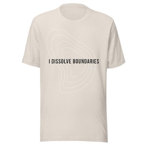 Pode incluir: Uma camiseta bege clara com o texto preto "I DISSOLVE BOUNDARIES". Acima do texto, há um design abstrato branco de linhas curvas concêntricas. A camiseta tem mangas curtas e gola redonda.