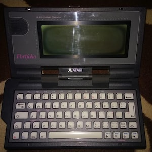 Könnte beinhalten: Ein Vintage Atari Portfolio Handheld-Computer. Das Gerät ist dunkelgrau mit einem kleinen Bildschirm und einer vollständigen Tastatur. Das Wort "Portfolio" ist in Rosa links oben aufgedruckt. Das Atari-Logo ist über der Tastatur sichtbar.