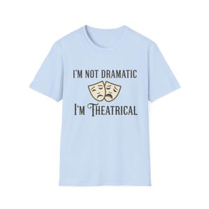 May include: Light blue t-shirt with the text "I'M NOT DRAMATIC I'M THEATRICAL" in black font. Above the text is a graphic of two theater masks, one smiling and one frowning, in a tan color. The shirt has short sleeves and a crew neck.