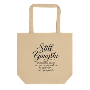 Op de afbeelding: Een beige draagtas met twee schouderbanden voorzien van zwarte tekst die luidt Still Gangsta I whisper to myself as I put on my readers to apply my estrogen patch. Het ontwerp is gecentreerd op de voorzijde van de rechthoekige stoffen tas.