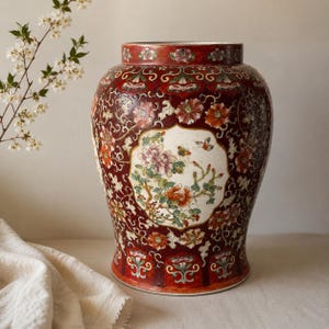 May include: A large ceramic jar with a deep red background featuring intricate white floral scrollwork and a central cream-colored cartouche. The cartouche displays a detailed painting of colorful peonies, green foliage, and small flying butterflies.